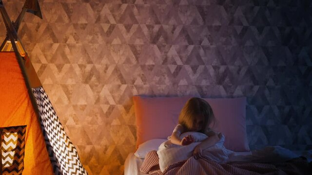Nervous little girl scratches pink plaid holding soft pillow on bed near illuminated by night lamp play wigwam in semidark bedroom at home slow motion