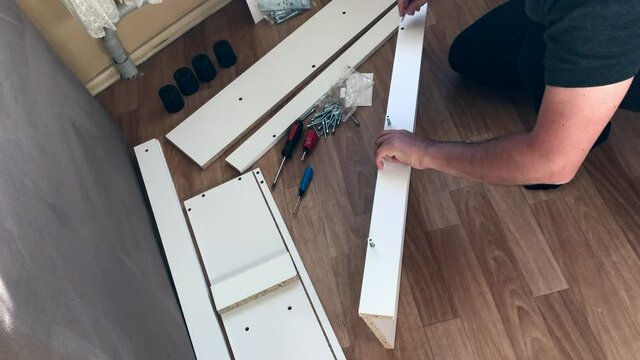 The man collects furniture at home. It connects furniture elements with each other with a confirmat screws. Other parts are spread out on the floor.