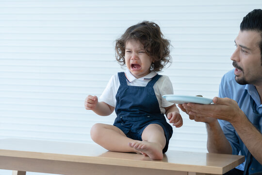 Little Caucasian Kid Girl With Curly Hair Is Sitting On Table Crying. Her Lips Stained With Tomato Sauce And Young Single Dad Feeding His Child Meal At Home