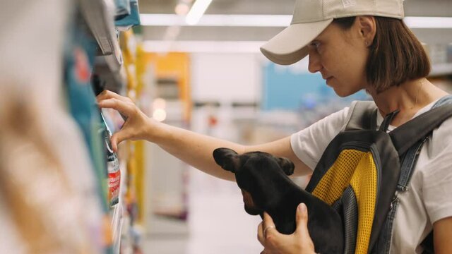 Young woman shopping at the supermarket with little toy terrier dog in her hands. She choosing dog food for her pet. High quality 4k footage