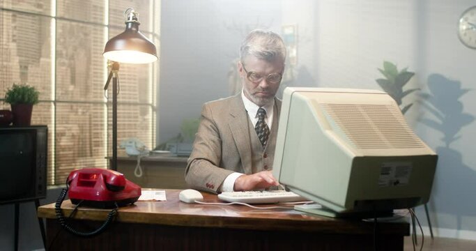 Caucasian male in glasses sitting at the desk in retro room and working on computer. Man programist typing on keyboard and looking seriously. Vintage style of 90's