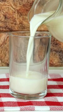Fresh natural milk is poured into glass from pitcher against brick wall background on in cage napkin. Filling drinking glass with milk. Healthy dairy product food concept. Vertical video.