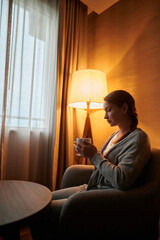 A young woman is sitting in a room in an armchair with a cup of tea in her hands. A cozy evening, a...