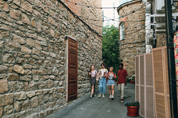 Group of young multiracial friends with drinks moving down narrow road between buildings