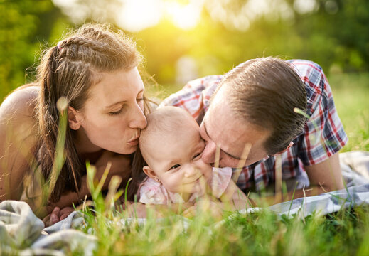 Happy family laying on a picnic blanket in nature with their baby in the middle and kissing her face together - Parenthood and childhood family concept outdoors in a park
