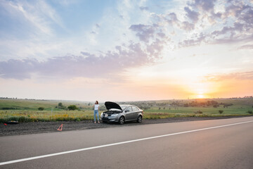 A young girl stands near a broken-down car in the middle of the highway during sunset and tries to call for help on the phone. Breakdown and repair of the car. Waiting for help.