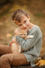  Boy with red cat ones autumn garden background close up photo. Kid with ginger cat on a autumn background. Beautiful autumn portrait of boy with ginger cat in the hands.