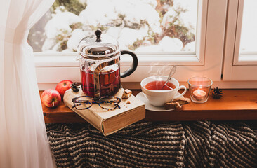 Winter relax at cozy home. Glass teapot with hot tea and book by the window.