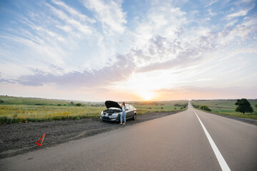 A young girl stands near a broken-down car in the middle of the highway during sunset and tries to...