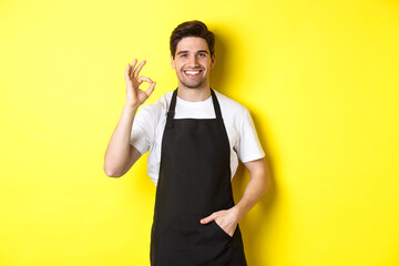 Young smiling barista in black apron showing okay sign, recommending coffee shop or restaurant,...