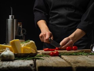 The chef is cutting tomatoes on a wooden cutting board. Nearby lies cheese, herbs, and other ingredients. Cooking salad, pie, pizza toppings. Wooden texture, black background. Restaurant, hotel, cafe.