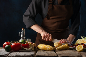 The chef slices the corn into pieces to prepare a traditional American dish - boiled corn with vegetables, lemon and crayfish. There are many ingredients on the wooden table. Restaurant, hotel.