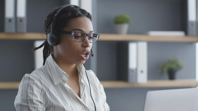 Young confident african american woman call center operator wearing headset talking with client via video chat on laptop