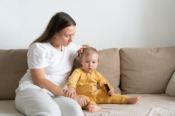 Horizontal portrait of young mother in casual sitting on sofa with adorable blond blue-eyed girl and grooming baby hair. Concept of mother care. Copy space