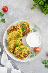 Vegetable zucchini pancakes with cheese, dill and parsley on a plate with sour cream on a gray concrete background. Top view, copy space.