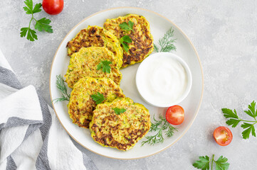 Vegetable zucchini pancakes with cheese, dill and parsley on a plate with sour cream on a gray concrete background. Top view, copy space.