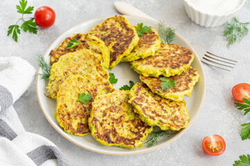 Vegetable zucchini pancakes with cheese, dill and parsley on a plate with sour cream on a gray concrete background. Top view, copy space.