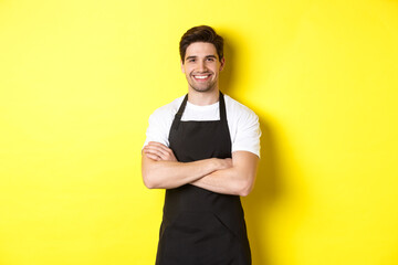 Smiling male waiter in black apron standing confident, cross arms on chest against yellow background