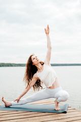 Fototapeta na wymiar Beautiful young woman in white sports clothes in warrior yoga pose on wooden pierce on lake outdoor.