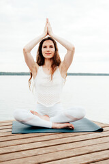 Fototapeta na wymiar Beautiful young woman in white sports clothes is sitting on wooden pier on lake in lotus position with namaste gesture.