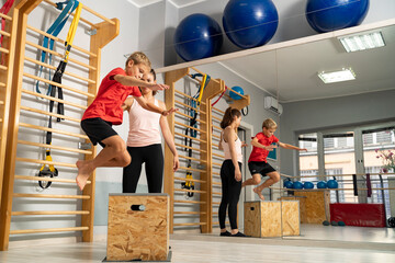 Energetic kid jumping on wooden box in the gym with help of his coach, teacher 