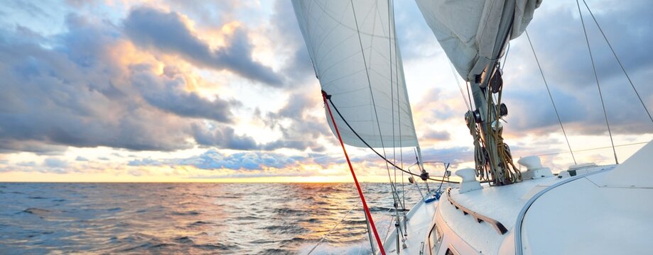 Yacht sailing in an open sea at sunset. Close-up view of the deck, mast and sails. Clear sky after the rain, dramatic glowing clouds, golden sunlight, waves and water splashes, cyclone. Epic seascape