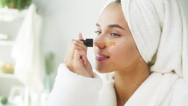 Woman applying face serum using pipette for moisturizing, morning skincare routine. Young female student wearing bathrobe looking in mirror in spa salon. Natural day make up concept.