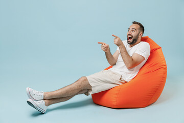 Full length young surprised excited happy man in white t-shirt sit in bag chair point index finger...