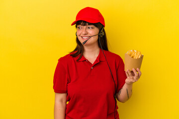 Young curvy caucasian woman fast food restaurant worker holding fries isolated on blue background looks aside smiling, cheerful and pleasant.