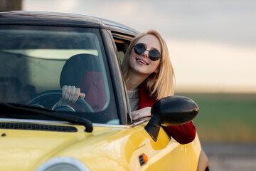 Style blonde woman in sunglasses and red coat in car at countryside
