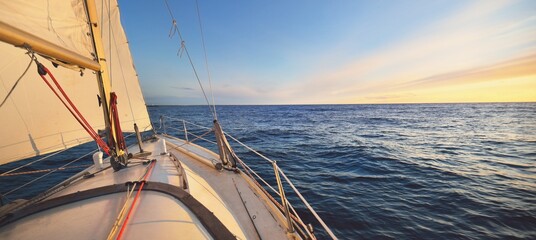 Yacht sailing in an open sea at sunset. Close-up view of the deck, mast and sails. Clear sky after the rain, glowing clouds, golden sunlight. Panoramic seascape