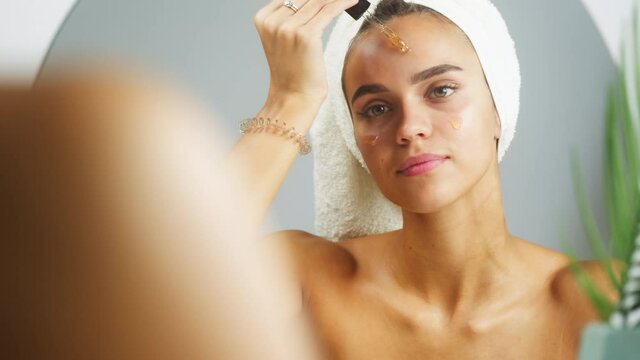 Woman applying face serum using pipette for moisturizing, morning skincare routine. Young female student looking in mirror in bathroom or spa with bare shoulders. Natural day make up concept.