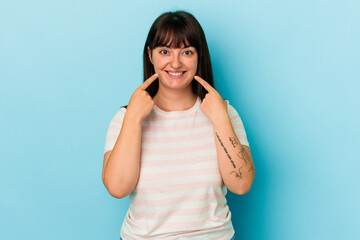 Young curvy caucasian woman isolated on blue background smiles, pointing fingers at mouth.