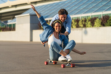 Man and woman have fun on longboard: cheerful male push joyful female sitting on skateboard and laughing. Happy couple enjoy summer time outdoors in city urban skate park. Relationship and lifestyle © DimaBerlin