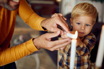 Little boy and his father lighting menorah candles while celebrating Hanukkah at home. - obrazy, fototapety, plakaty