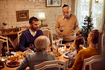Mature Jewish man holds a toast during Hanukkah family meal at dining table.
