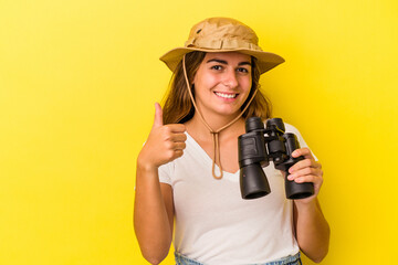 Young caucasian woman holding binoculars isolated on yellow background  smiling and raising thumb up