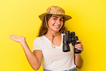 Young caucasian woman holding binoculars isolated on yellow background  showing a copy space on a palm and holding another hand on waist.