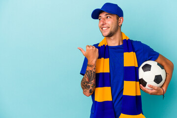 Young soccer fan man holding a ball isolated on blue background  points with thumb finger away, laughing and carefree.
