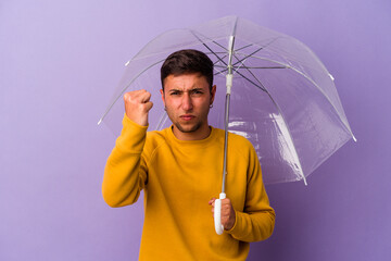 Young caucasian man holding umbrella isolated on purple background  showing fist to camera, aggressive facial expression.