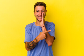 Young caucasian man with tattoos isolated on yellow background  saying a gossip, pointing to side...