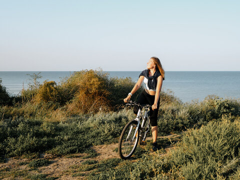 Young sports girl with a white bike on the seashore on a summer morning at sunrise. Outdoor sports training. Tinted image.