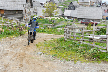 Motorcyclist is racing off- road motorcycle in wilderness surrounded with log cabins in green environment.