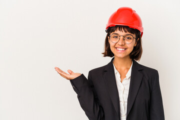 Young architect woman with red helmet isolated on white background showing a copy space on a palm and holding another hand on waist.