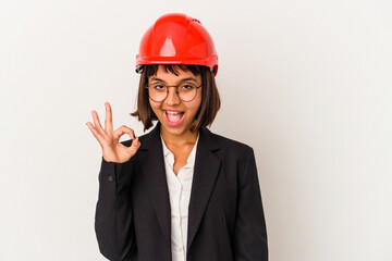 Young architect woman with red helmet isolated on white background winks an eye and holds an okay gesture with hand.
