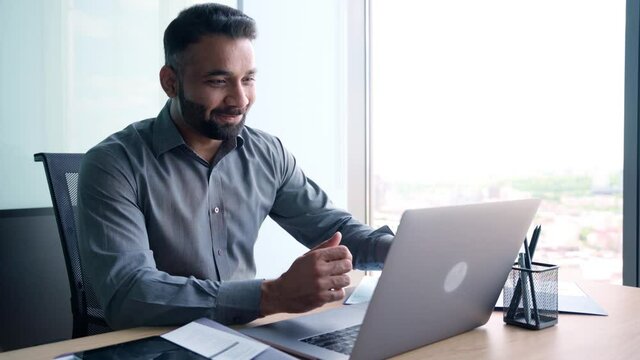 Successful happy smiling indian businessman sitting at desk having working videoconference virtual chat with client using pc laptop computer in contemporary corporation office. Business concept.