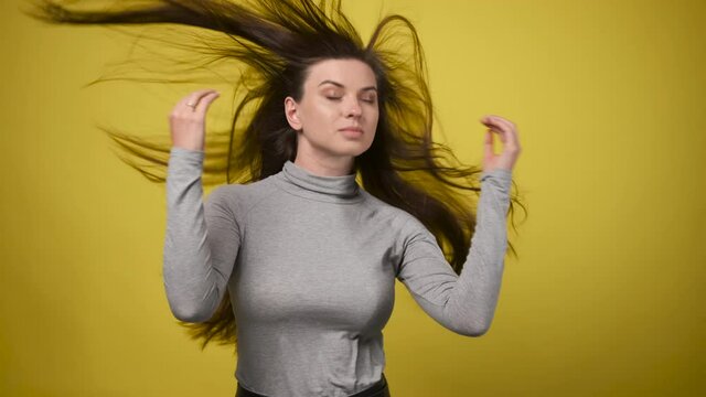 slender woman in gray turtleneck stands against yellow background in studio and straightens her hair, which is fluttering in strong wind.