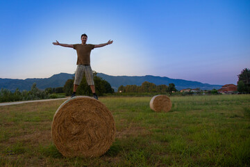 Naklejka na ściany i meble smiling happy man on haystack with arms outstretched in summer before sunset