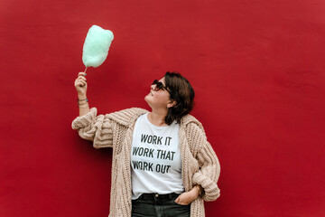 Image of a woman looking at a cotton candy wearing dark sweater glasses with one hand in her pocket relaxed