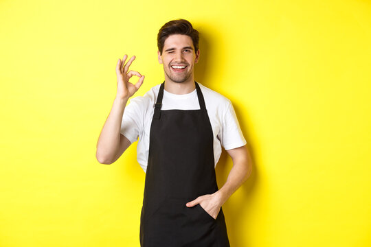 Confident and handsome waiter showing ok sign, wearing black apron and standing against yellow background